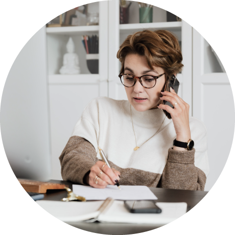 A woman with glasses talking on the phone while writing on a piece of paper at her desk.